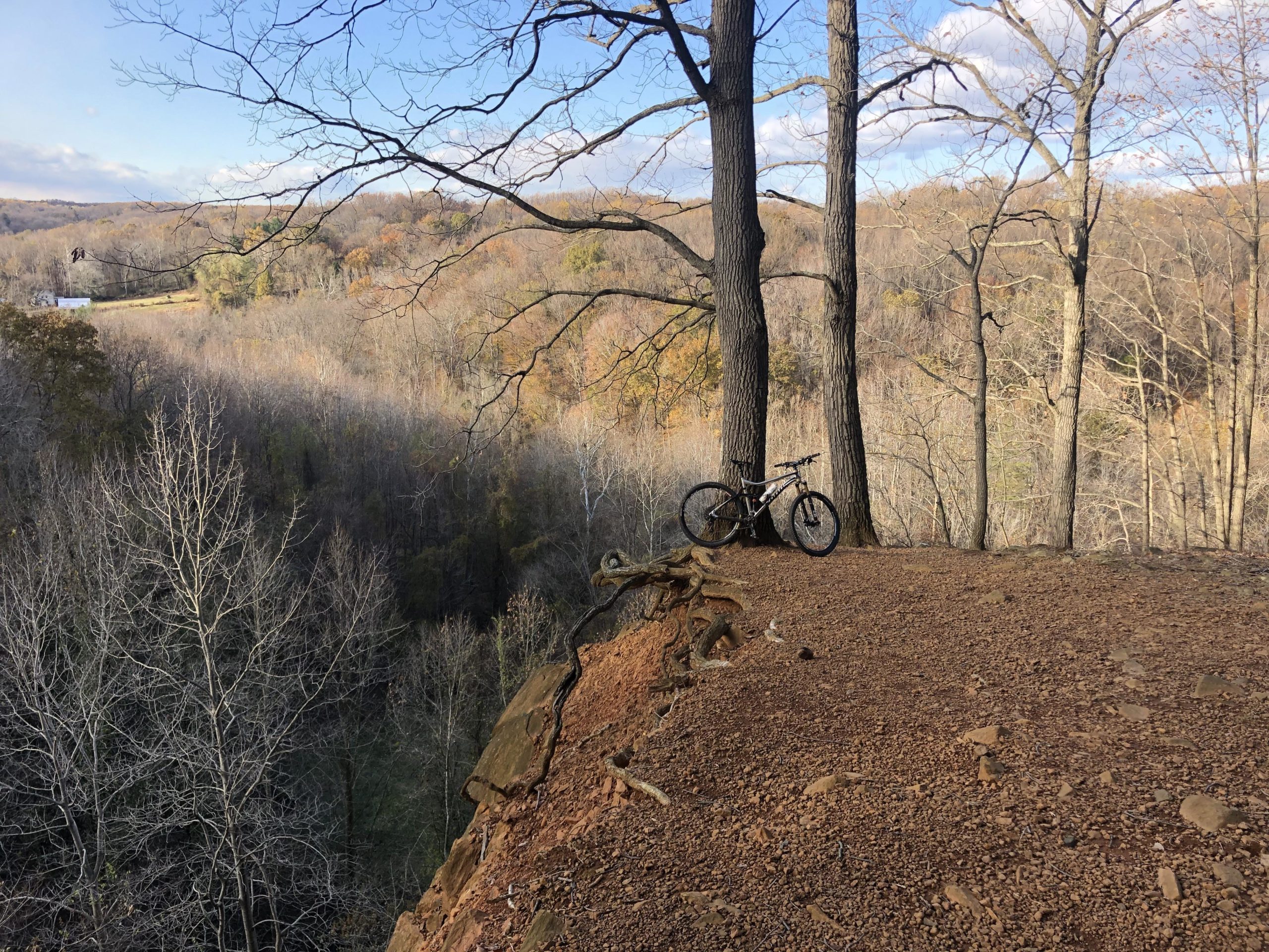 A mountain bike rests against two tall trees at the edge of a rocky overlook, with a scenic view of a forested valley below. The landscape features a mix of bare trees and autumn foliage, under a partly cloudy sky. Jerusalem Mill trail mountain bike trail.