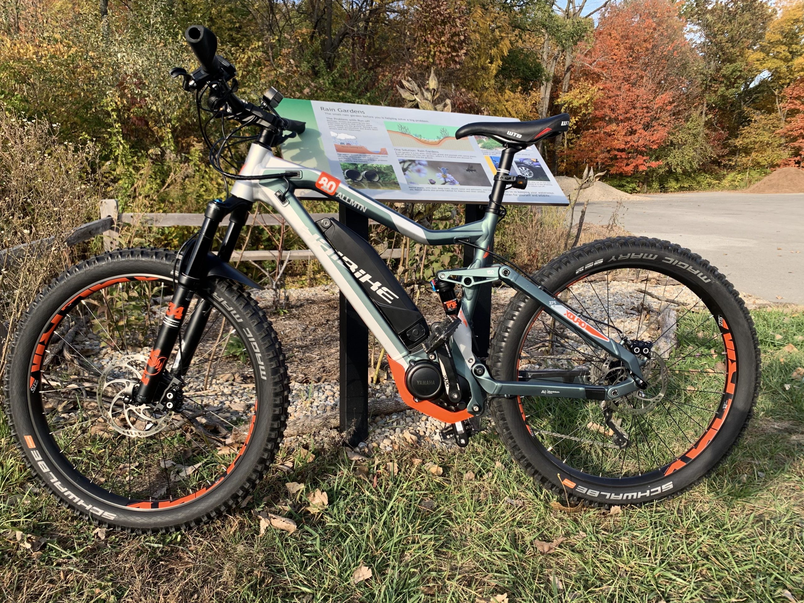 Electric mountain bike parked in front of an informational sign about rain gardens, surrounded by autumn foliage and grass. Fort Harrison State Park mountain bike trail.