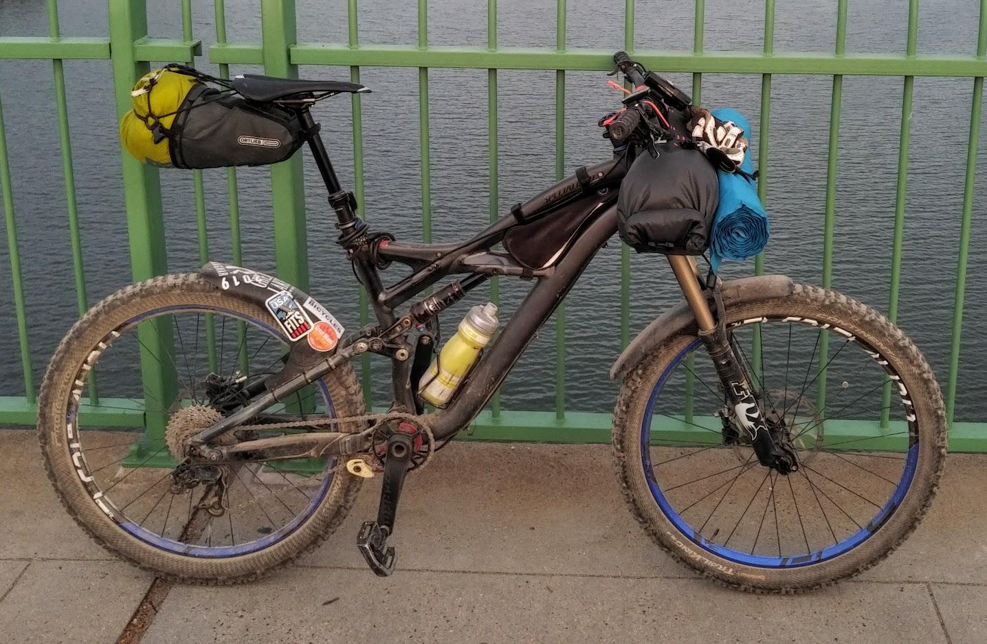 Specialized Enduro Expert: A black mountain bike with visible dirt and mud, equipped with various bags and a rolled mat on the handlebars, parked beside a green railing overlooking a body of water. The bike features blue accents on the wheels and several stickers on the frame.