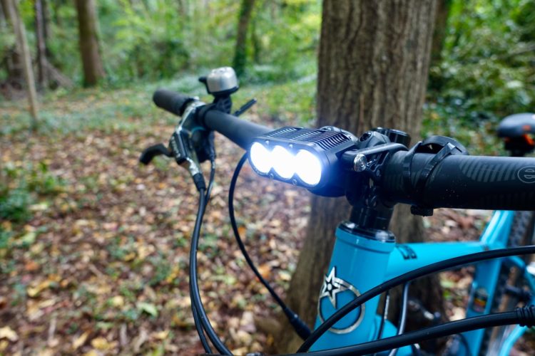 Close-up view of a bicycle handlebar featuring a bright LED headlight, set in a natural environment with trees and fallen leaves in the background. The handlebars are black, while the bike frame is a vibrant blue.