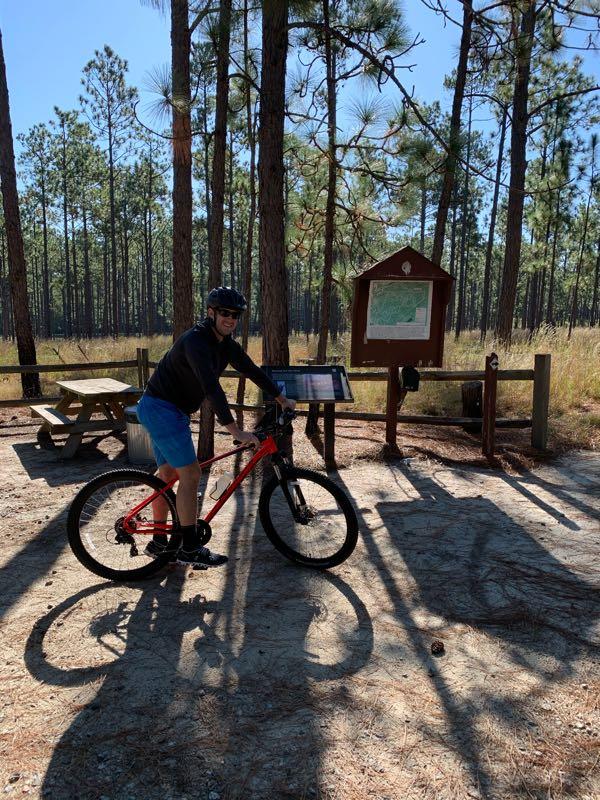 A person wearing a helmet and cycling gear is standing next to a red mountain bike at a trailhead. They are positioned beside an informational sign in a forested area with tall pine trees and a picnic table nearby. Sunlight filters through the trees, casting shadows on the ground. Cheraw State Park Mountain Bike Trail mountain bike trail.