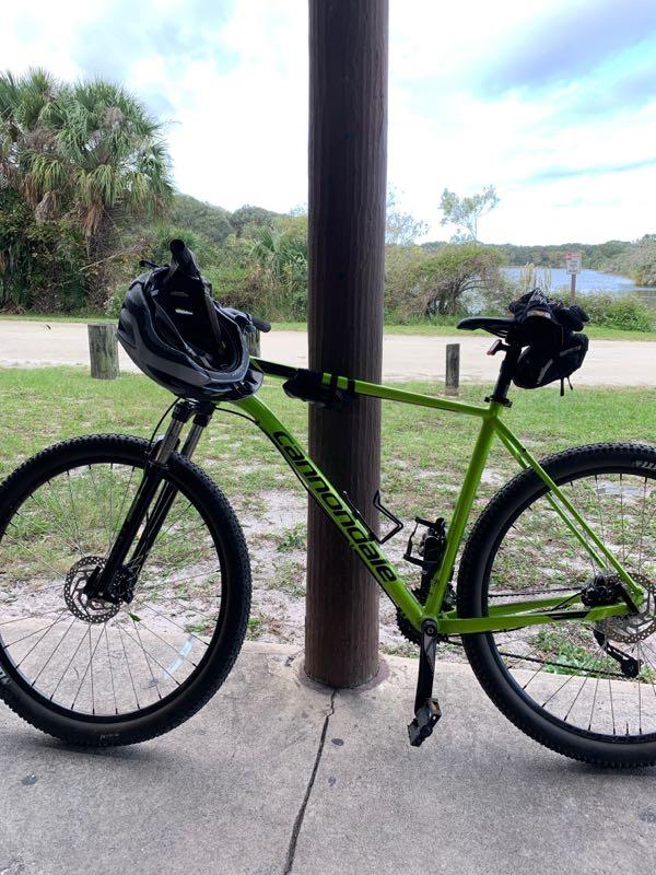 A green Cannondale mountain bike is parked under a wooden structure, with a black helmet placed on the handlebars. In the background, there are palm trees and a view of a path leading to a lake. The sky is partly cloudy. Kathryn Abby Hanna Park mountain bike trail.