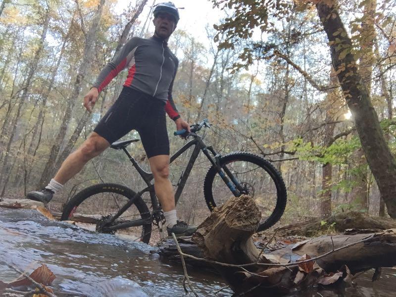 A cyclist standing on a log over a stream, wearing a helmet and cycling gear, with a mountain bike beside him. The background features a wooded area with autumn foliage and soft sunlight filtering through the trees. Hard Labor Creek State Park mountain bike trail.