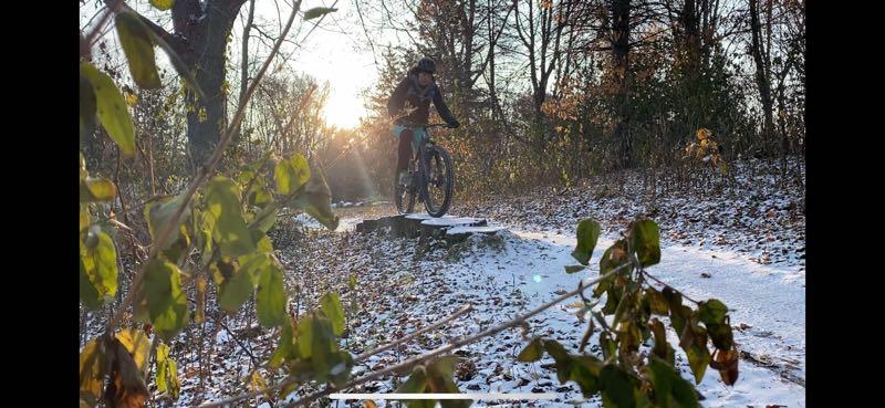 A person riding a mountain bike over a small wooden bridge on a snow-covered path surrounded by trees during sunset. The scene captures autumn leaves and a warm glow from the setting sun. Elm Creek Park mountain bike trail.