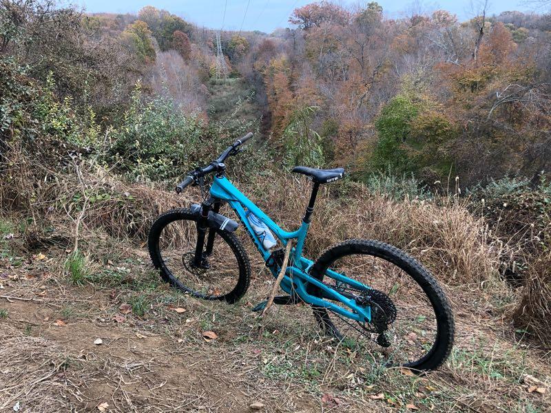 A turquoise mountain bike rests on a dirt path surrounded by tall grass and shrubs, overlooking a scenic view of a valley with autumn foliage in shades of orange and green. Power lines stretch across the landscape in the background. Patapsco Valley State Park (Avalon Area) mountain bike trail.