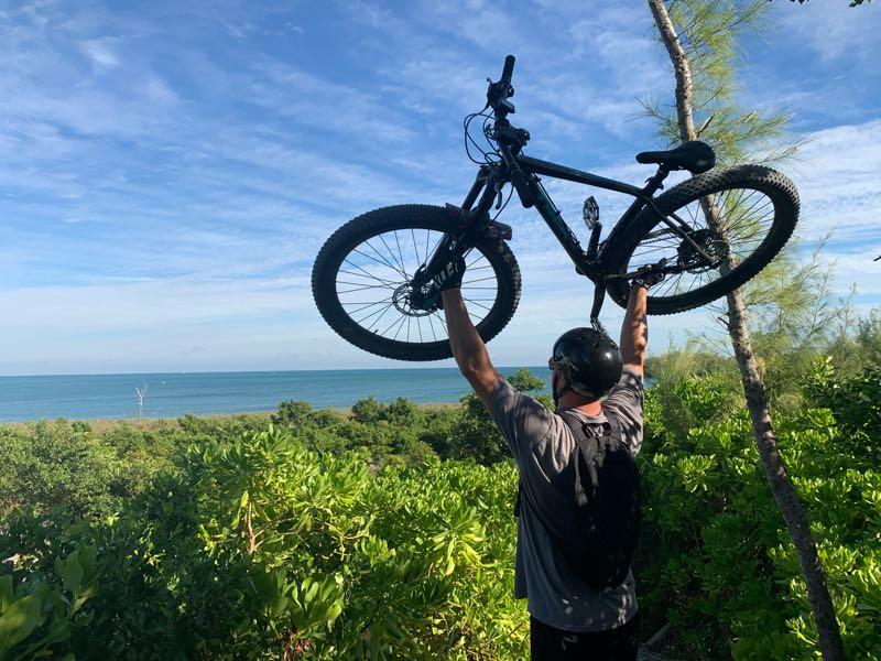 A man wearing a helmet and backpack holds a mountain bike above his head while standing on a hill overlooking a scenic view of the ocean and greenery under a clear blue sky. Virginia Key North Point mountain bike trail.