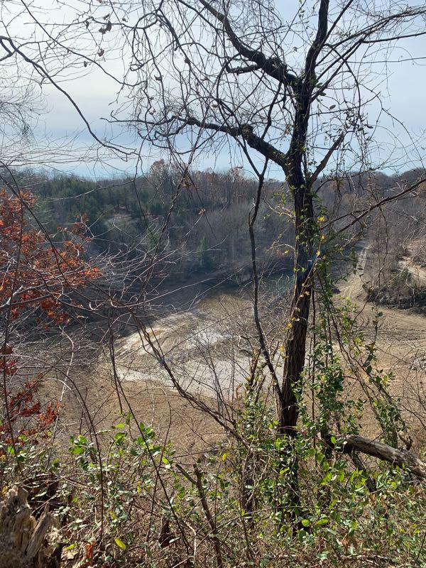 A scenic view from a hillside, showcasing a barren landscape with a winding riverbed partially visible. The foreground features a tree with sparse branches, while patches of greenery and dried leaves surround the area. In the background, rolling hills are visible under a cloudy sky, indicating a tranquil and natural setting. Idlewood mountain bike trail.