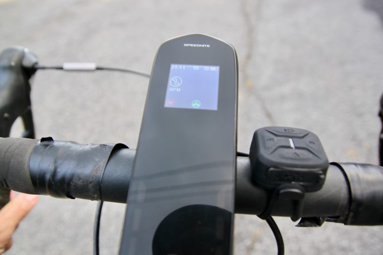 Close-up of a bicycle handlebar featuring a digital display showing RPM and other metrics, with a control button nearby, and a partially wrapped handle with black tape. The background is a blurred outdoor scene.