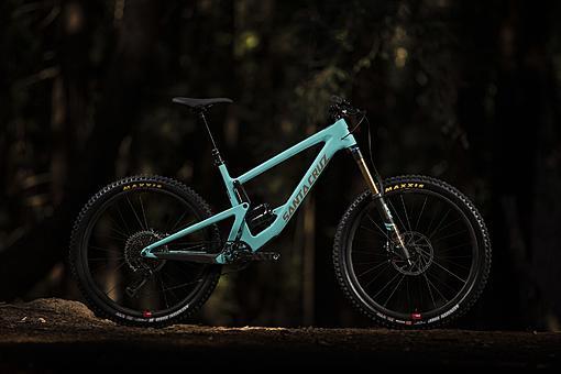 Santa Cruz Bronson: A turquoise mountain bike is positioned on a forest floor, surrounded by dark, natural lighting that highlights its sleek design and features.