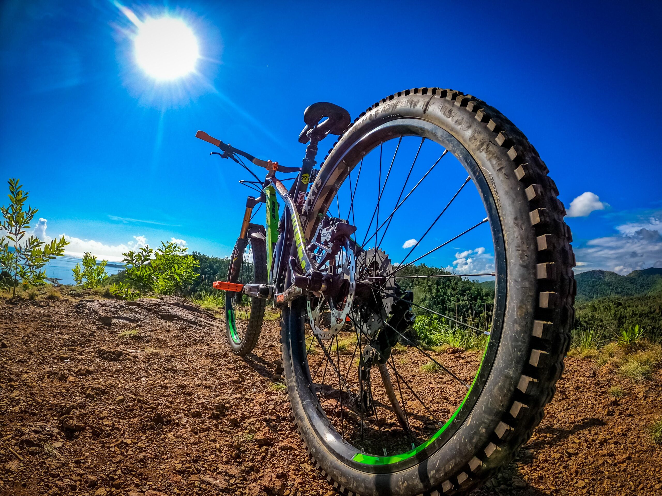 Mondraker Dune R: A close-up view of a mountain bike parked on a dirt path, with the sun shining brightly in a clear blue sky. The bike's front tire is prominently featured, showcasing its tread pattern, while the surrounding landscape includes green foliage and distant hills.