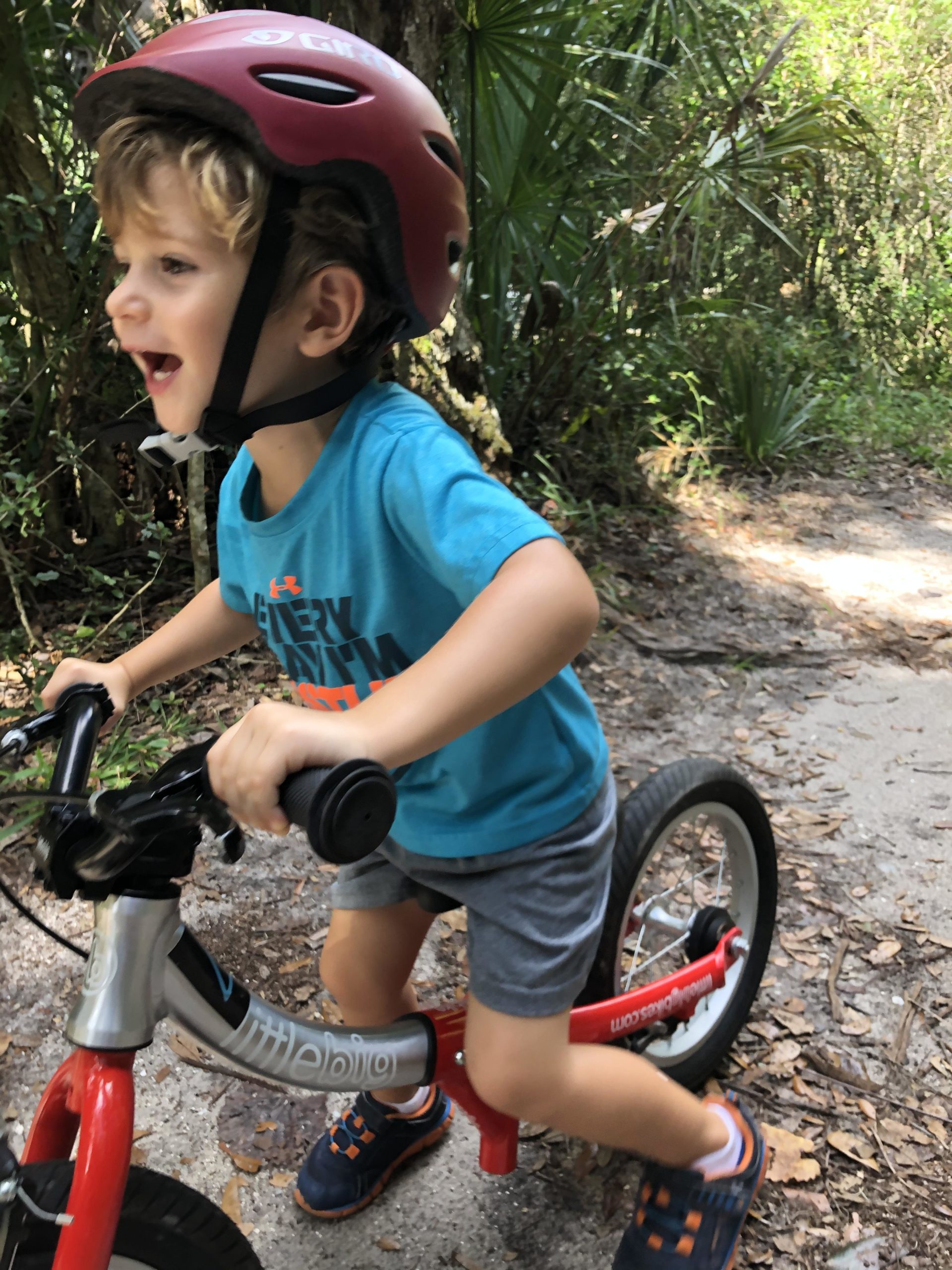 A young child wearing a red helmet rides a balance bike along a dirt path in a lush, green wooded area. The child has curly hair and is smiling joyfully as they pedal, wearing a blue t-shirt and gray shorts. Kathryn Abby Hanna Park mountain bike trail.