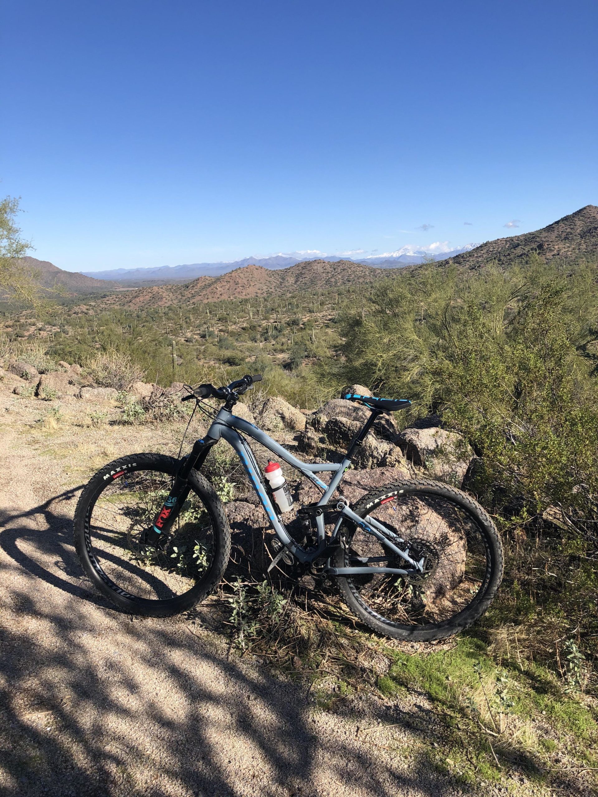 Mountain bike resting on rocks with a scenic view of desert hills and blue sky in the background. The landscape features green shrubs and cacti, showcasing a rugged outdoor environment. Hawes Loop mountain bike trail.