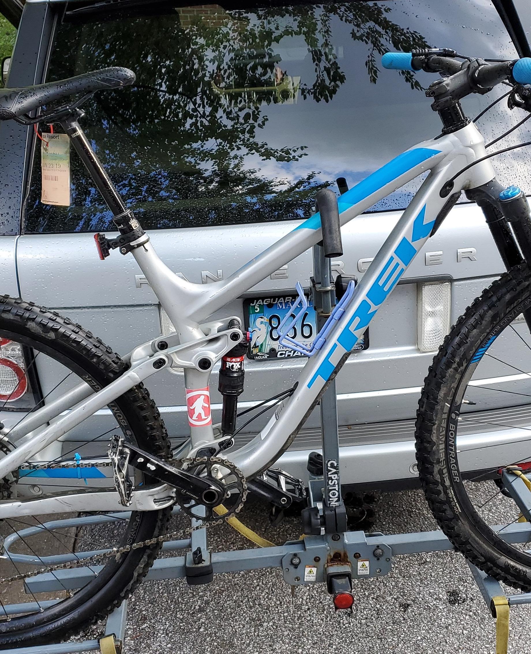 Trek Fuel EX 9 29: A gray and blue Trek mountain bike secured on a bike rack attached to a silver SUV. The bike features a sleek frame, knobby tires, and a visible suspension system. A tag is hanging from the seat, and a sticker is on the bike's frame. The background includes reflections of trees and clouds on the vehicle's rear window.