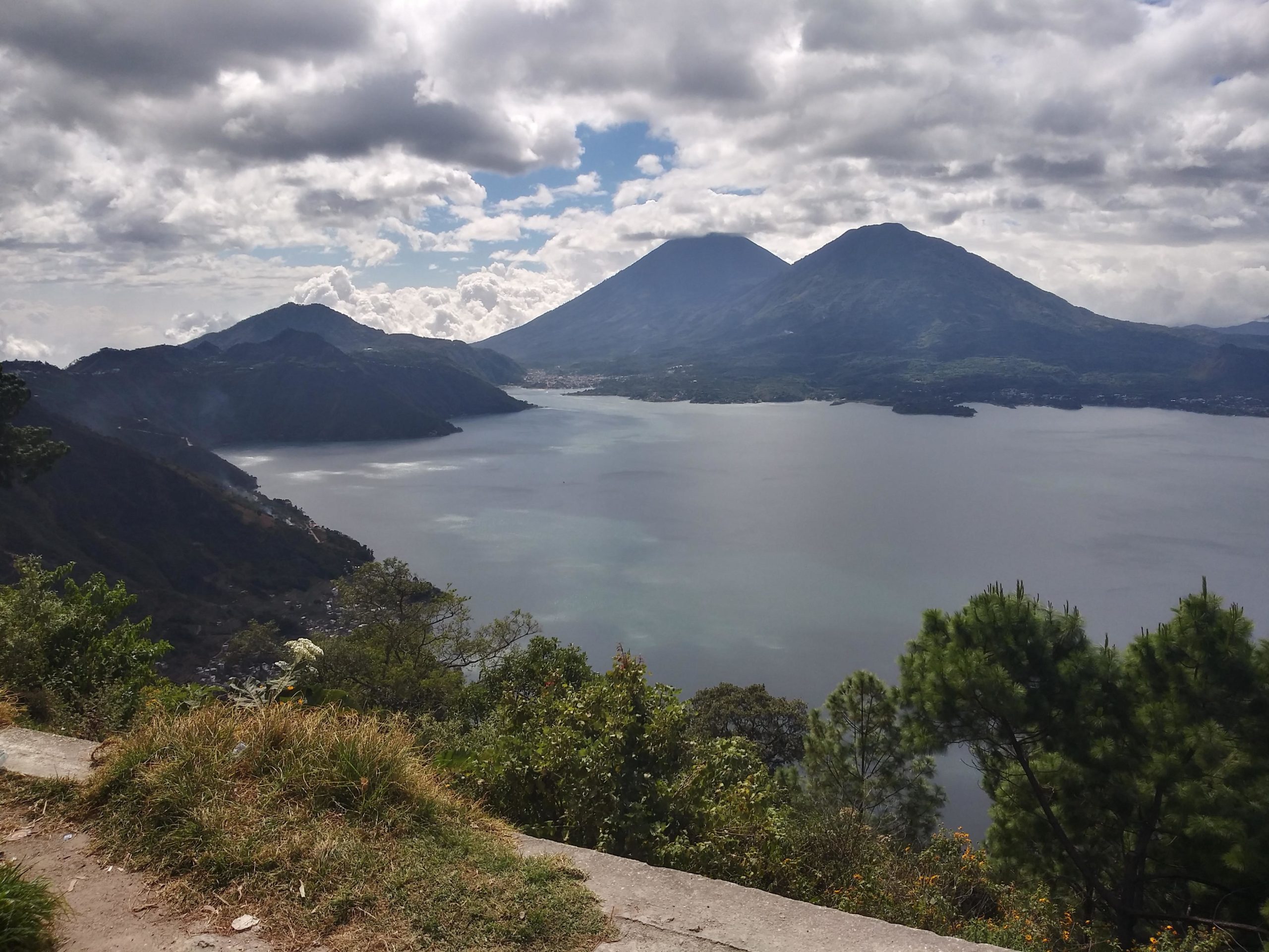 A panoramic view of a serene lake surrounded by lush mountains under a partially cloudy sky. The water reflects the clouds above, and the landscape features green vegetation along the shore, with two prominent volcanic peaks rising in the background. Pana to Pacaman Loop mountain bike trail.
