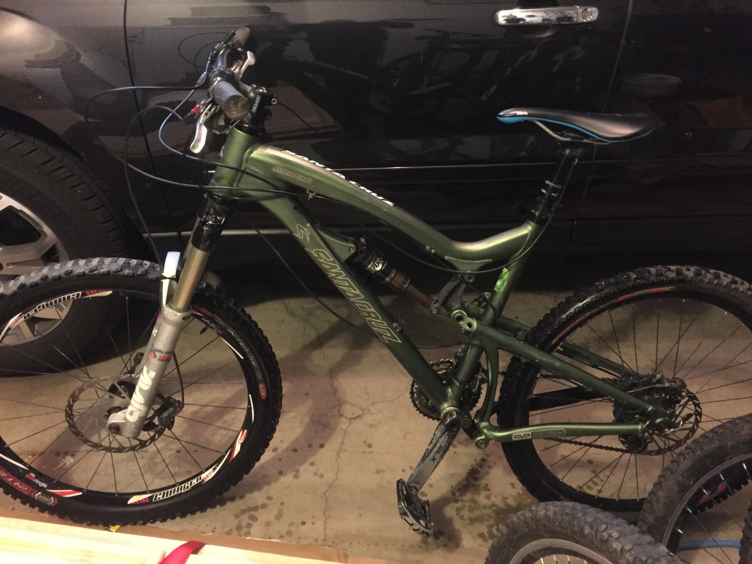 Santa Cruz Nomad: A green mountain bike with a silver front suspension fork, disc brakes, and knobby tires, resting in a garage beside a vehicle. The bike features a sleek design with prominent branding and is positioned on a concrete floor.