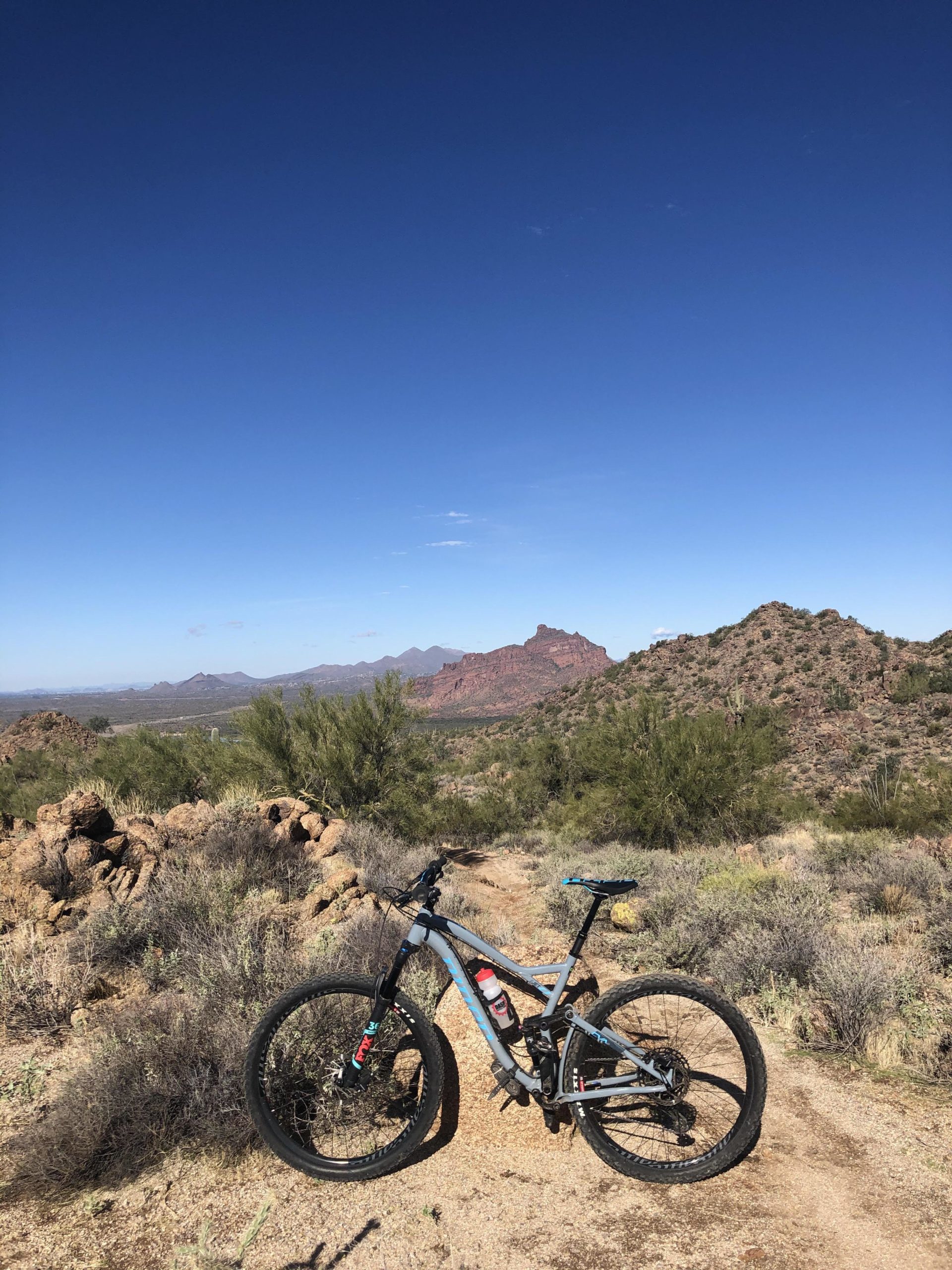 A mountain bike stands on a dirt trail with a backdrop of rugged mountains and a clear blue sky. The surrounding landscape features rocky terrain and sparse vegetation, highlighting an outdoor adventure setting. Hawes Loop mountain bike trail.