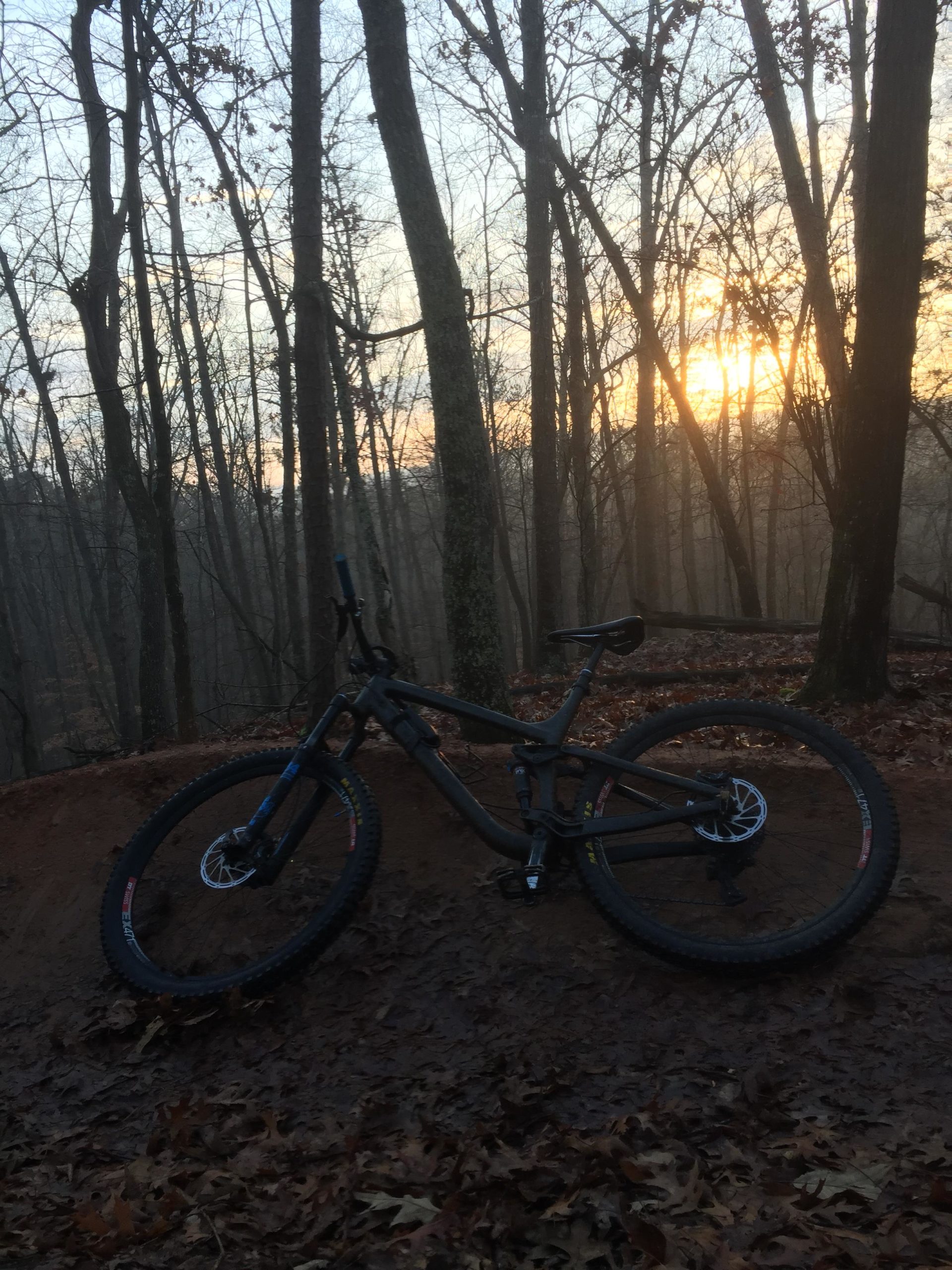 A mountain bike rests on a dirt mound surrounded by bare trees, with a hazy sunrise illuminating the background. Fallen leaves cover the ground, creating a tranquil atmosphere in a wooded area. Van Michael Trail mountain bike trail.