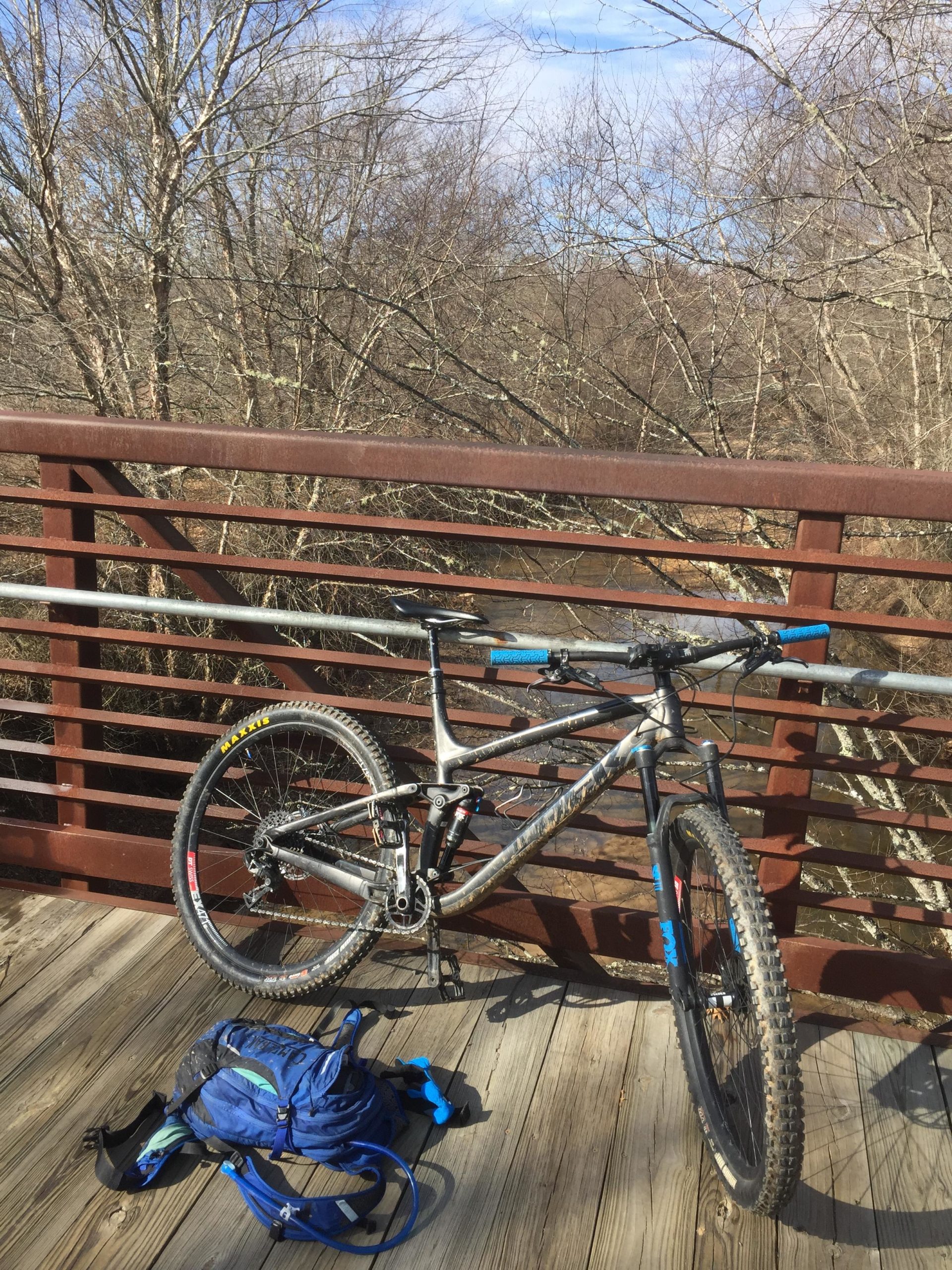 A mountain bike resting on a wooden bridge, with a blue backpack lying next to it. In the background, a tree-lined landscape and a river can be seen under a clear sky. Allatoona Creek Park mountain bike trail.