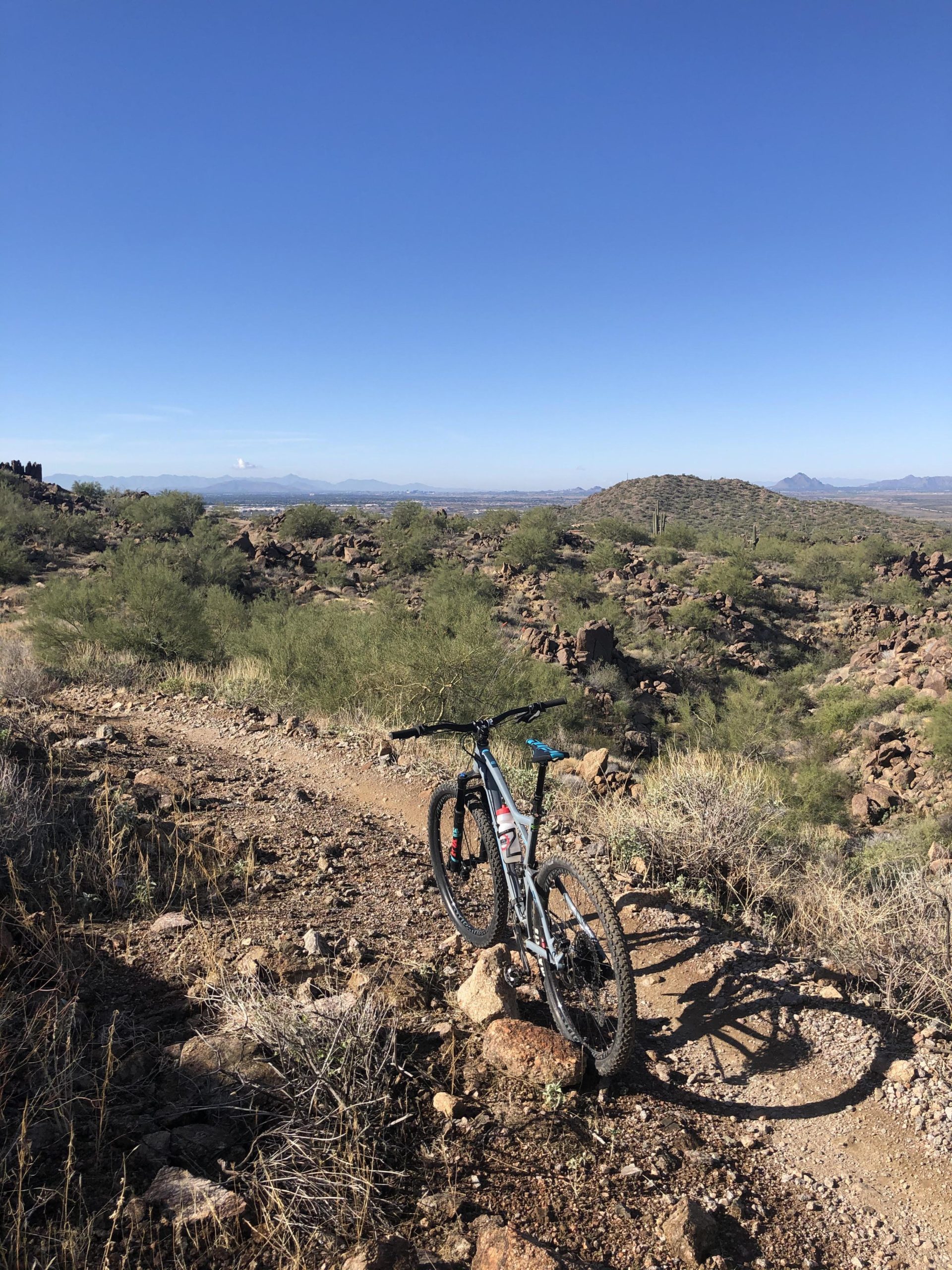 A mountain bike leaning against a rock on a dirt trail, surrounded by a desert landscape with scattered vegetation and rocky hills under a clear blue sky. Hawes Loop mountain bike trail.