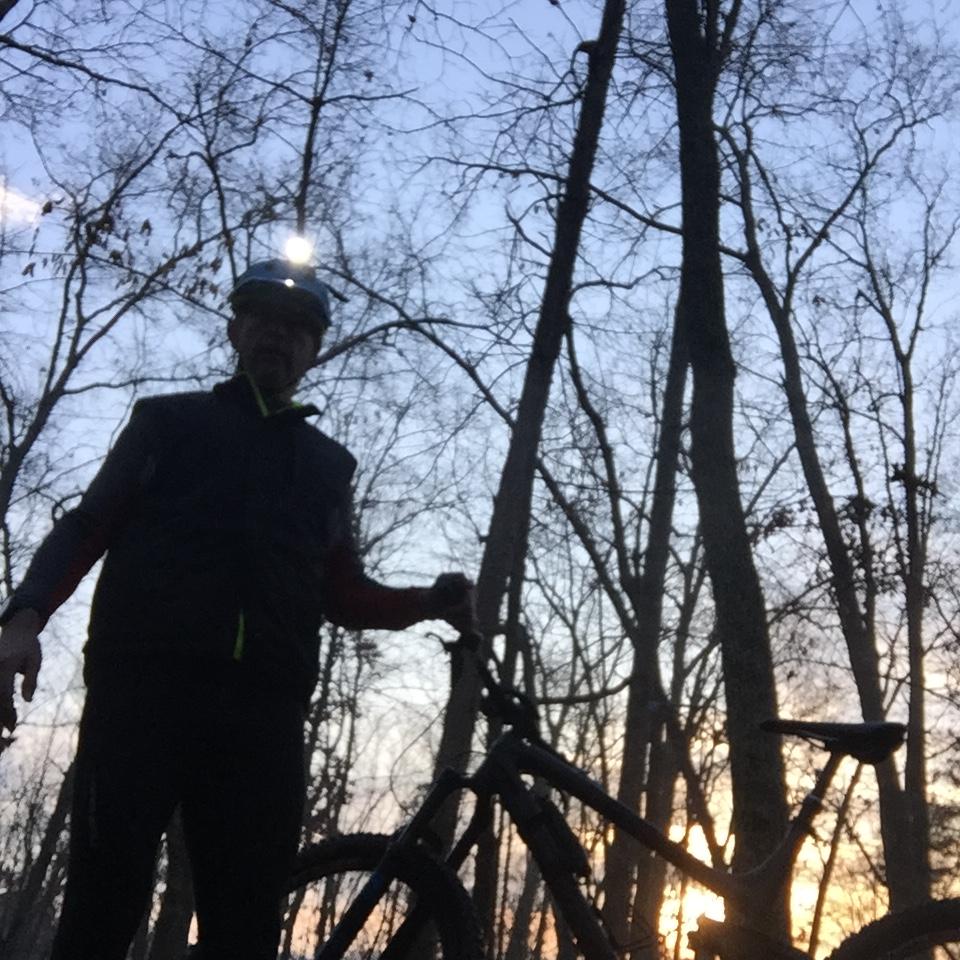 A person standing near a mountain bike in a forested area during sunset. The individual is wearing a helmet and a vest, with trees silhouetted against the twilight sky. The sun is setting in the background, creating a serene outdoor atmosphere. Van Michael Trail mountain bike trail.