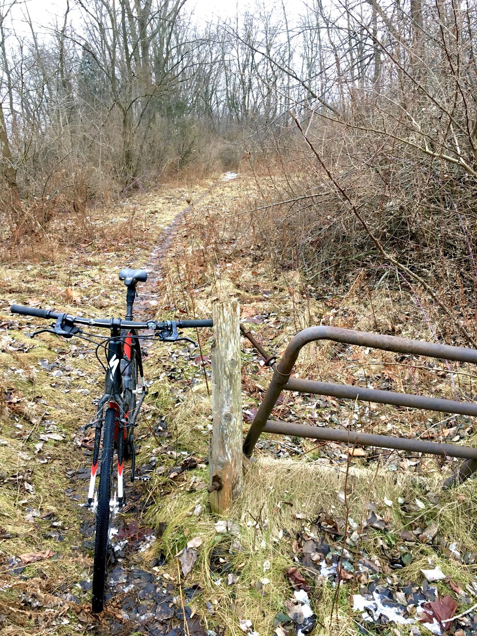 A mountain bike parked next to a wooden post and a metal gate, set along a narrow, overgrown trail in a wooded area. The scene features sparse vegetation, fallen leaves, and a hint of mud on the path, suggesting a chilly, possibly damp day. Hardy Rd. Trail mountain bike trail.