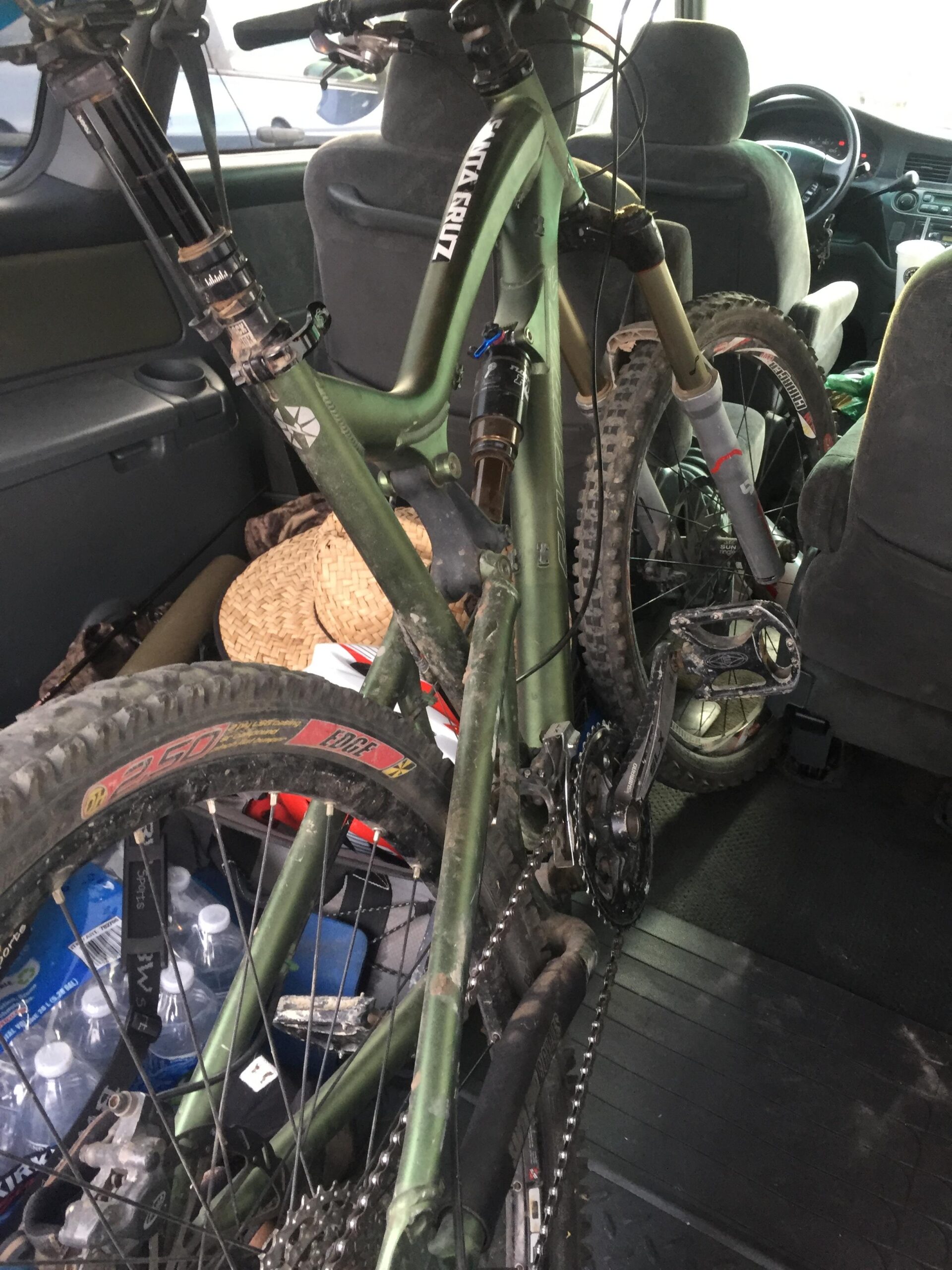 Santa Cruz Nomad: Alt text: A mountain bike with a green frame is positioned inside the back seat of a vehicle. The bike appears dusty and used, with visible dirt on the tires and components. Water bottles and other items are also visible in the background of the vehicle’s interior.