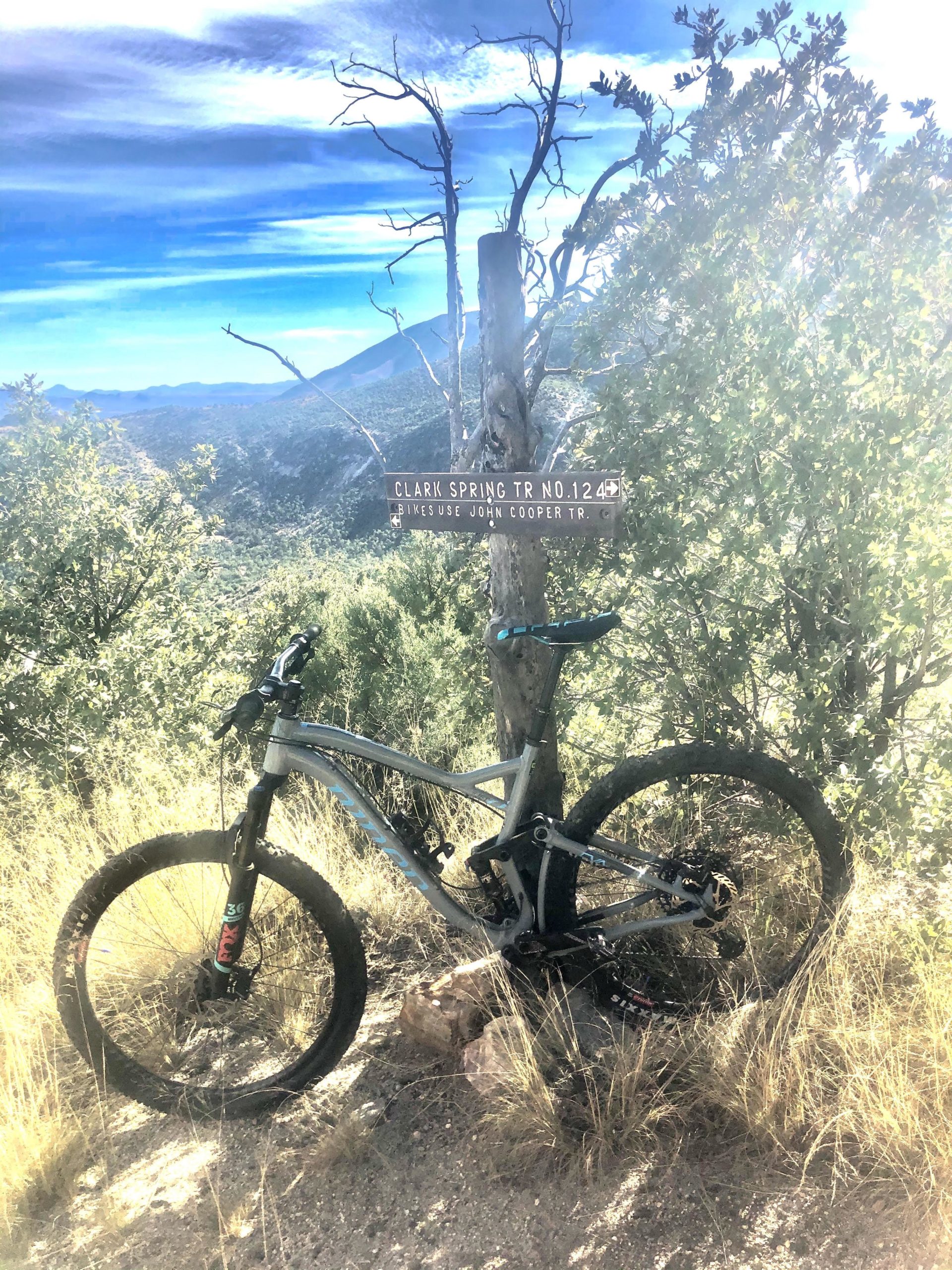 Mountain bike leaning against a wooden trail sign marking "Clark Spring Trail No. 124" in a sunlit landscape with tall grass and sparse trees. The background features a mountainous terrain and blue sky with wispy clouds. Perimeter Trail mountain bike trail.