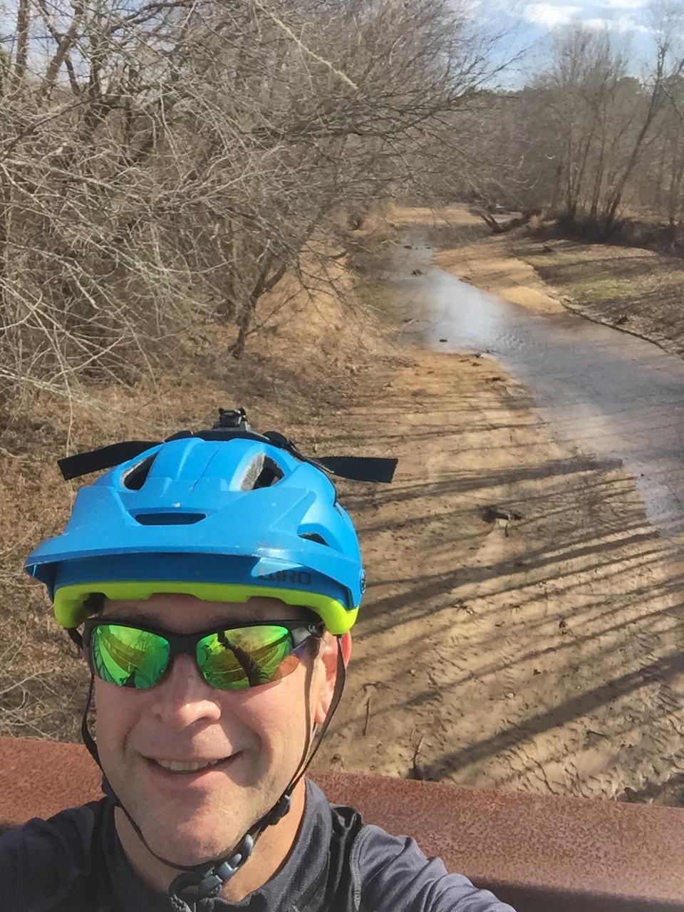 A person wearing a blue and green bicycle helmet and sunglasses smiles for a selfie while standing on a bridge overlooking a dry riverbed surrounded by bare trees. The scene is set under a clear sky with a few clouds. Allatoona Creek Park mountain bike trail.
