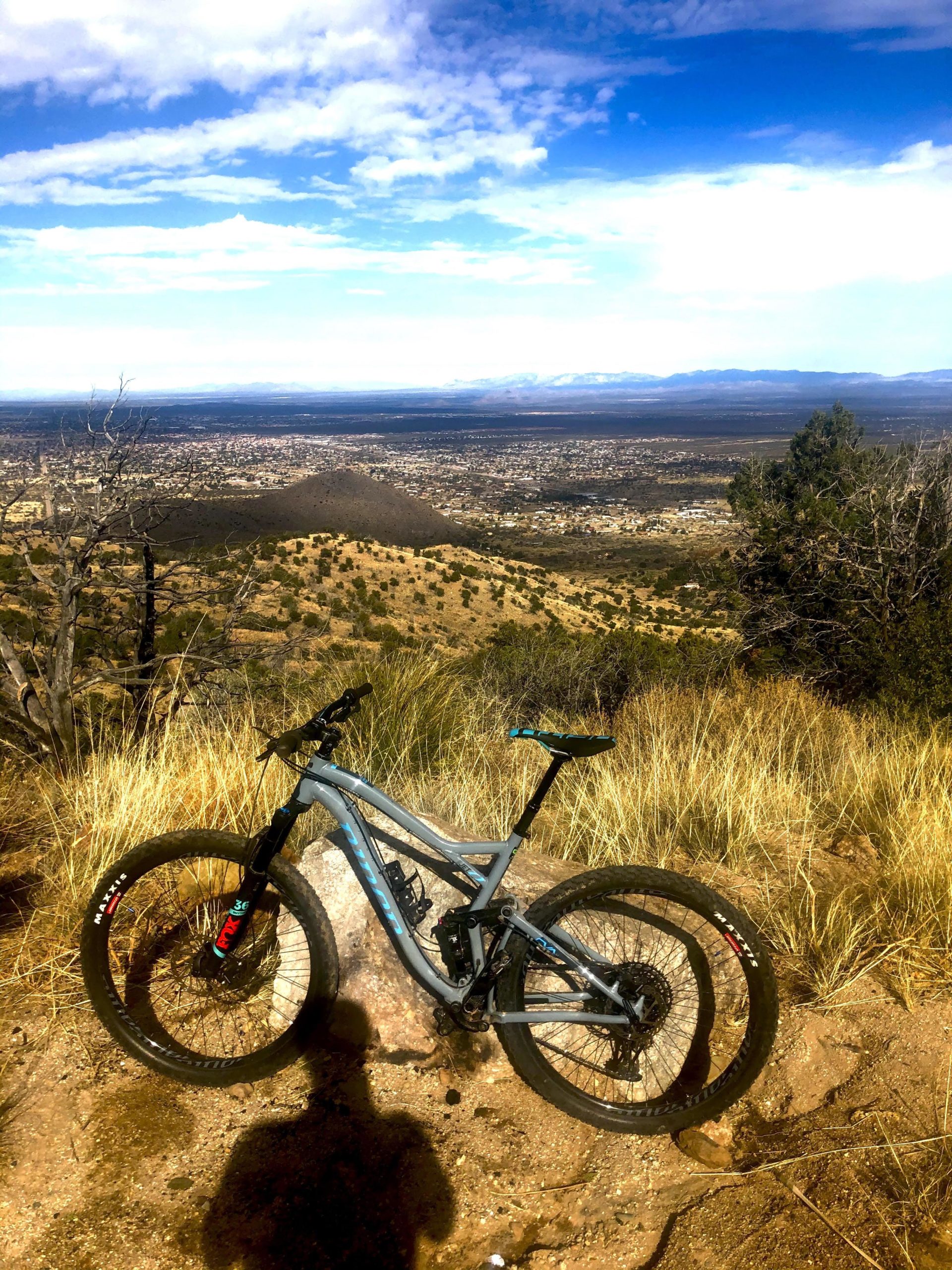 A mountain bike rests on a rocky outcrop with a scenic view of a valley and distant mountains. The landscape features dry grass, shrubs, and a clear blue sky with scattered clouds. The bike is positioned prominently in the foreground, casting a shadow on the ground. Perimeter Trail mountain bike trail.