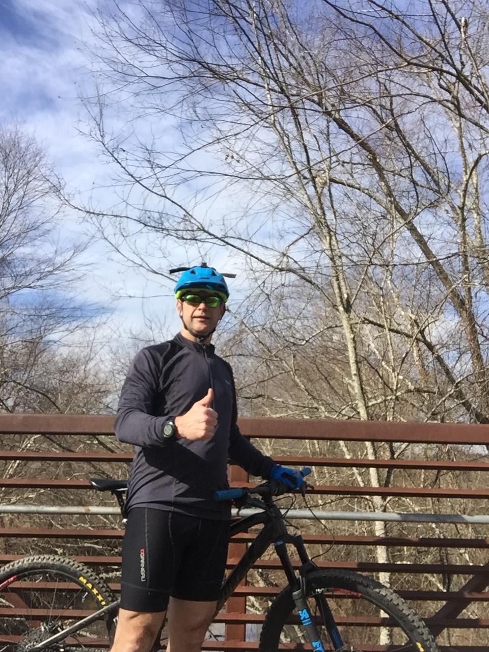 A person in athletic clothing and a blue helmet stands next to a mountain bike on a wooden bridge, giving a thumbs-up. Bare trees and a blue sky are visible in the background. Allatoona Creek Park mountain bike trail.