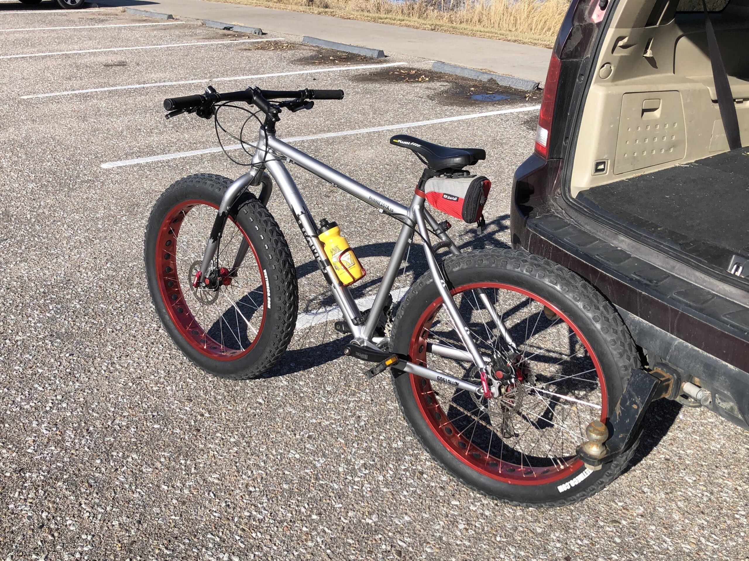 Framed Minnesota 2.0: A silver fat bike with red rims parked next to an open trunk of a vehicle in a gravel parking lot. A yellow water bottle is mounted on the bike frame, and a small black bag is attached to the bike's rear.