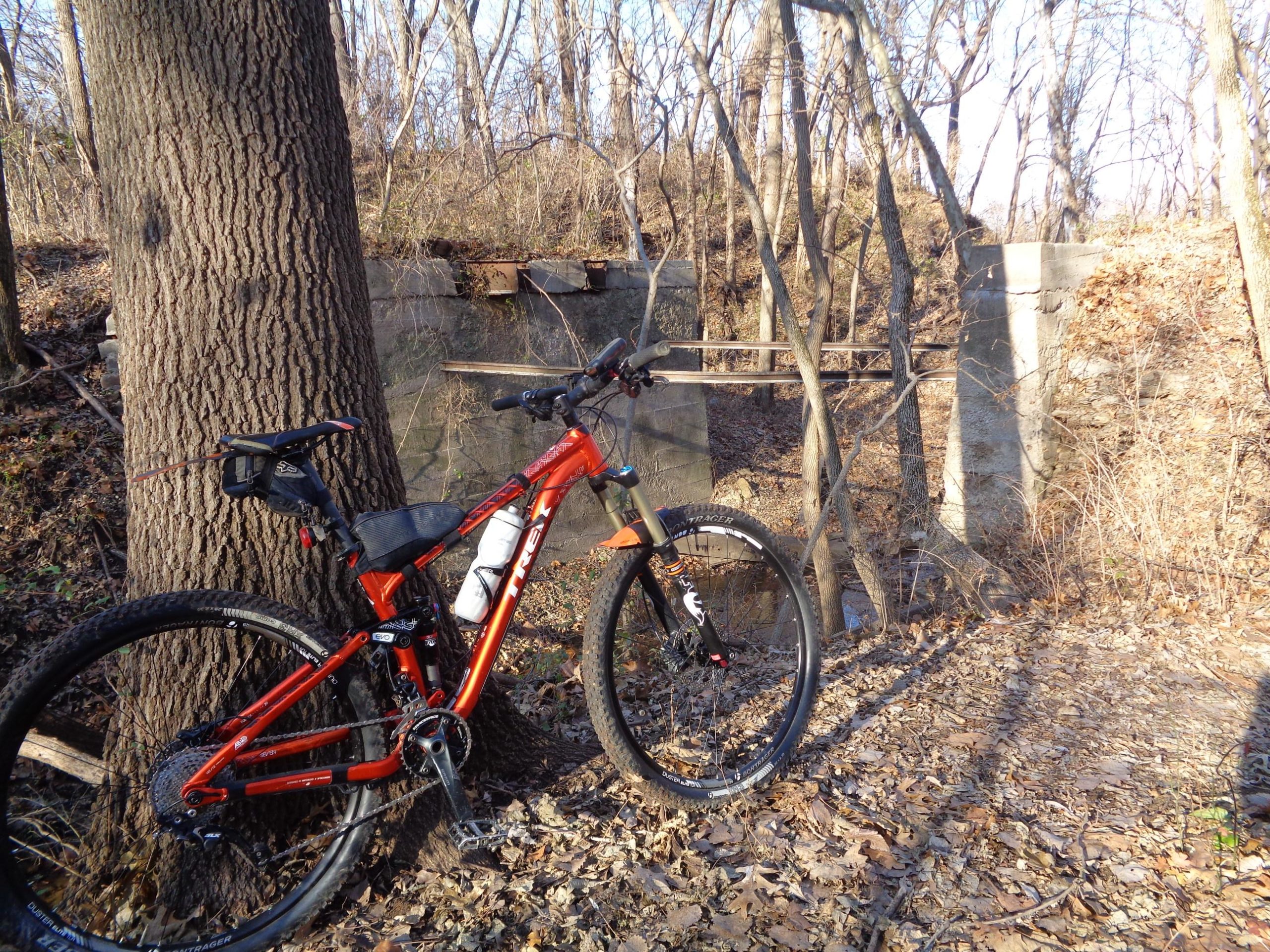 A vibrant orange mountain bike rests against a large tree in a wooded area, with fallen leaves scattered on the ground. In the background, remnants of an old structure, including concrete pillars and wooden beams, are visible among the trees. The scene is illuminated by soft sunlight filtering through the branches, creating a serene outdoor setting. Lehigh Portland Trails mountain bike trail.