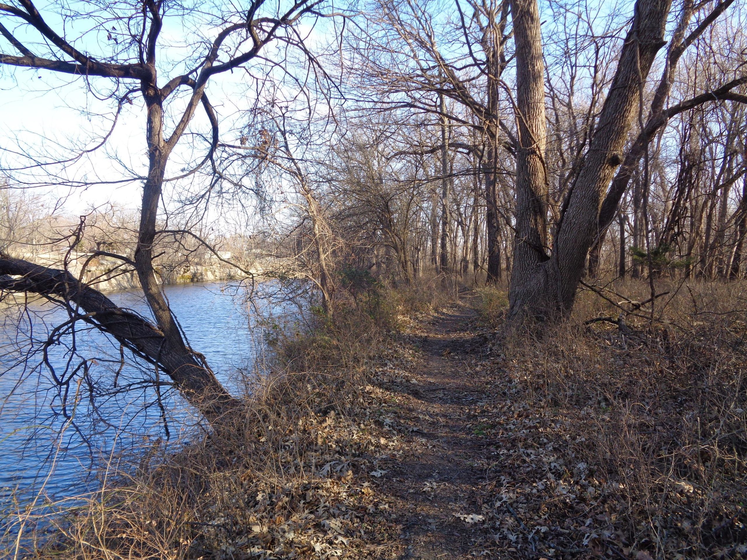 A narrow dirt path lined with bare trees and shrubs runs alongside a river. The landscape is mostly devoid of leaves, indicating a late autumn or early winter scene. The sky is clear and blue, casting a serene atmosphere over the tranquil water and surrounding nature. Lehigh Portland Trails mountain bike trail.