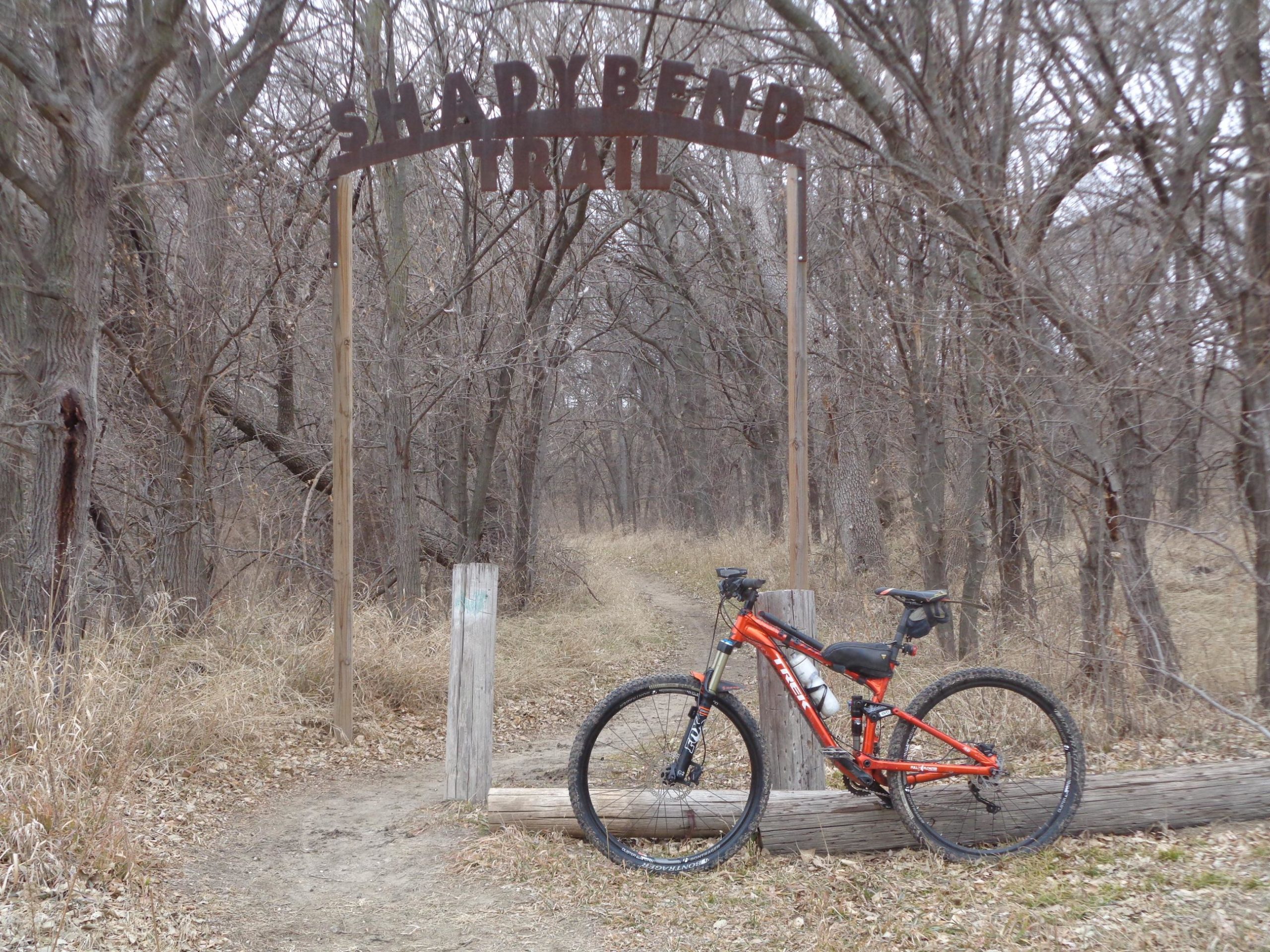 A mountain bike is leaning against a wooden post at the entrance of the Shadybend Trail, surrounded by bare trees and dry grass. The trail path is visible in the background, inviting exploration. Shady Bend Trail mountain bike trail.