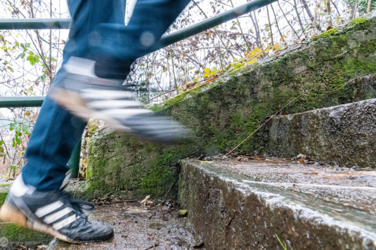 A close-up view of a person's foot in motion as they ascend wet, moss-covered outdoor steps, surrounded by foliage and greenery. The focus is on the foot wearing a black sneaker with white stripes, indicating movement and activity in a natural setting.