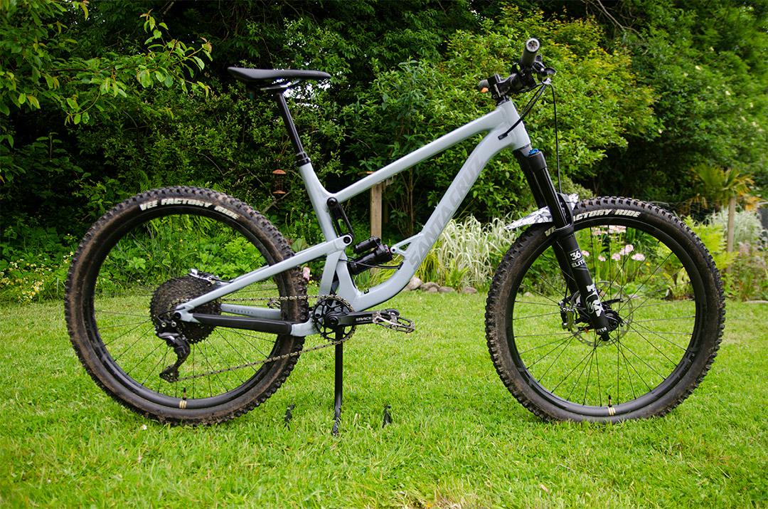 Santa Cruz Bronson 3 Alu: A side view of a modern mountain bike with a gray frame, featuring wide tires with knobby tread, a front suspension fork, and a rear shock. The bike is positioned on green grass with trees and shrubs in the background, suggesting an outdoor setting.