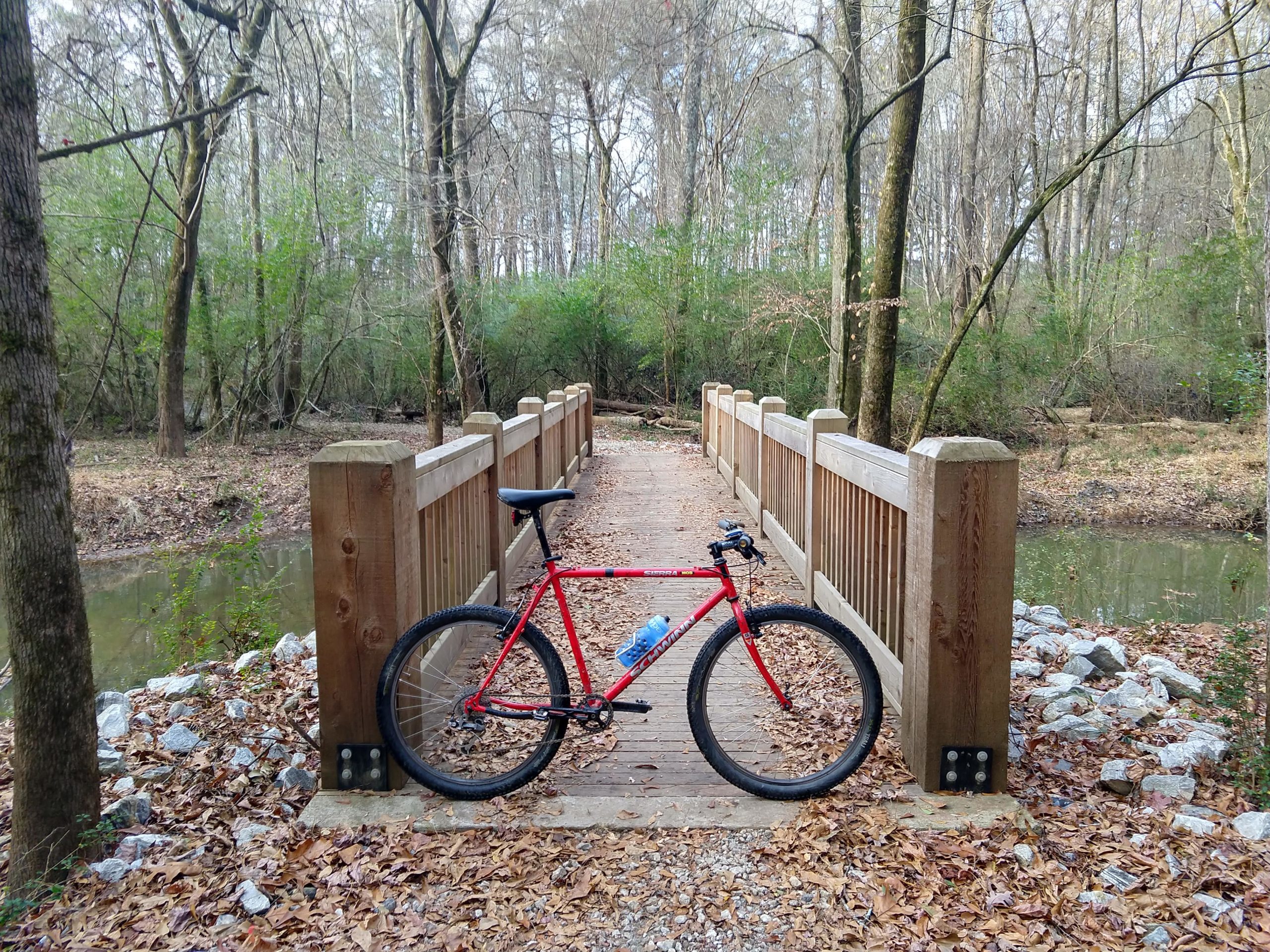 A red bicycle parked on a wooden bridge surrounded by trees and foliage, with a small stream underneath and leaves scattered on the ground. Southside Park mountain bike trail.