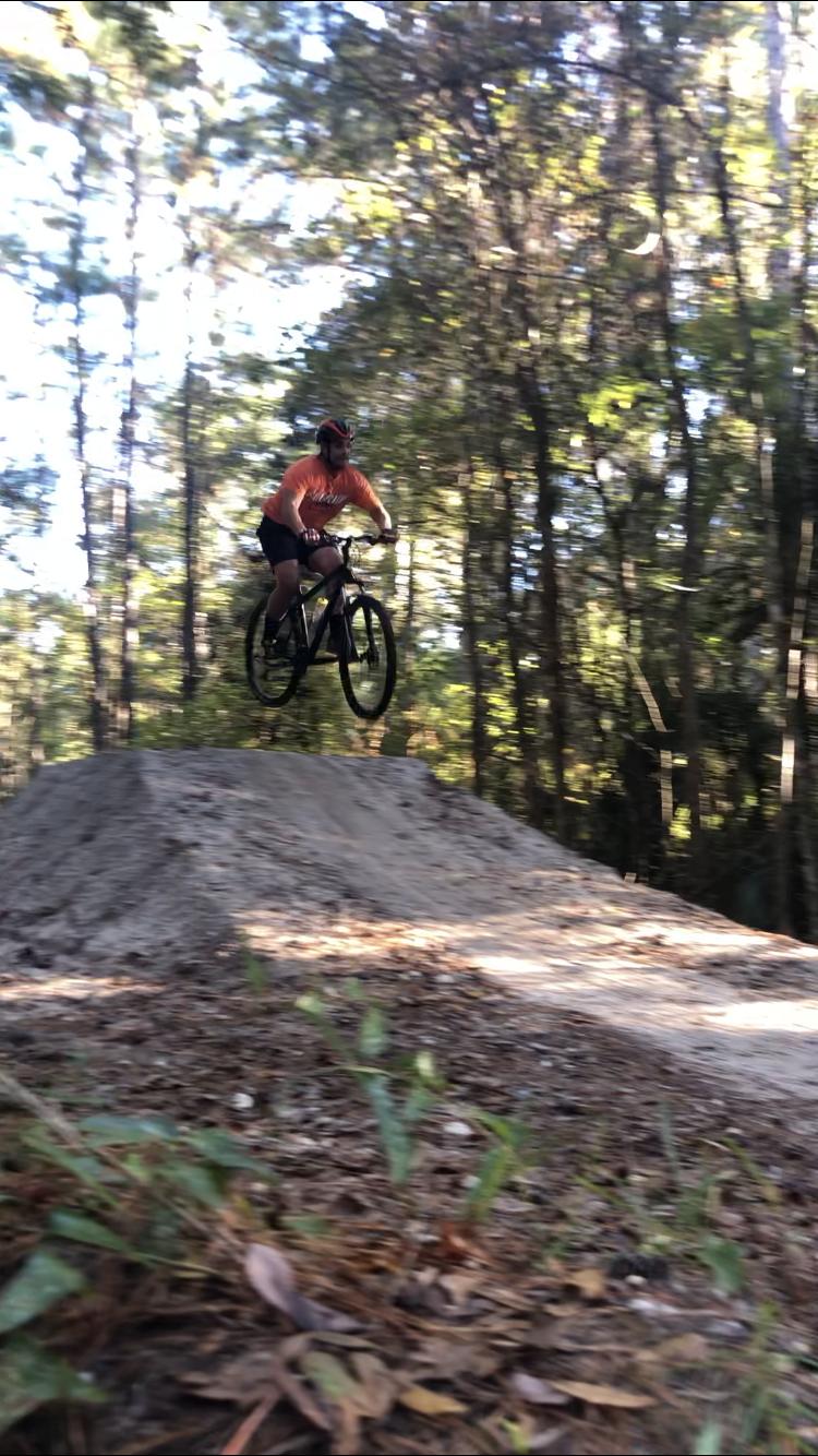 A person wearing an orange shirt and a helmet is jumping off a dirt jump on a mountain bike, surrounded by trees in a wooded area. The bike is airborne with the front wheel elevated as the rider descends from the jump. The scene captures motion, emphasizing the thrill of mountain biking. Tillie Fowler Regional Park mountain bike trail.