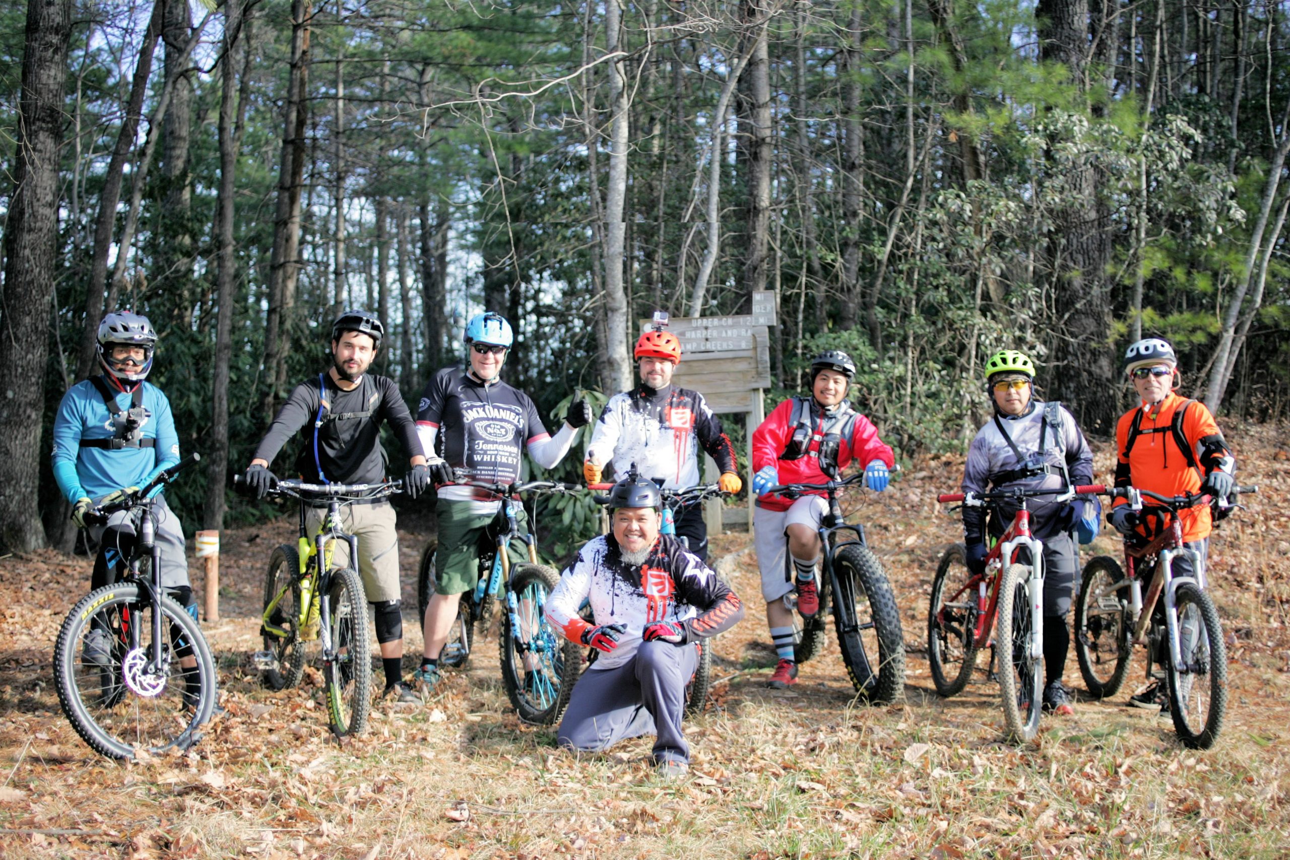 A group of eight mountain bikers posing for a photo in a wooded area. They are wearing cycling gear and helmets, with their bikes beside them. The backdrop features trees and a wooden sign partially visible in the background. The terrain is covered in fallen leaves, suggesting an autumn setting. Greentown Trail (268) mountain bike trail.