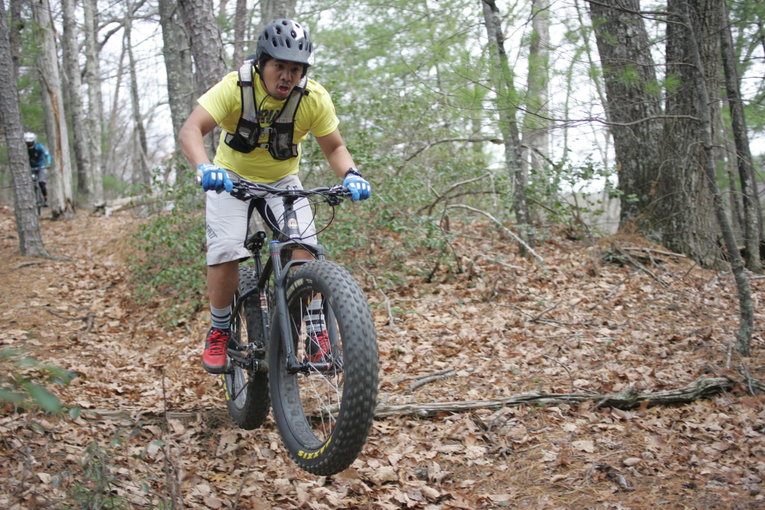 A cyclist in a yellow shirt and helmet performs a jump on a fat bike on a wooded trail covered with leaves and twigs. The background features trees and greenery, with another cyclist visible in the distance. Sinkhole mountain bike trail.
