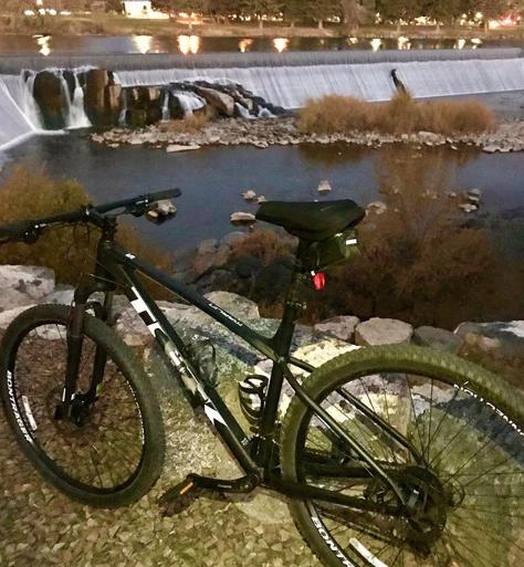 Trek Marlin 5: A black mountain bike parked near a flowing river with a waterfall, surrounded by rocks and shrubs. The scene is set during dusk, with soft lighting from nearby sources reflecting on the water.