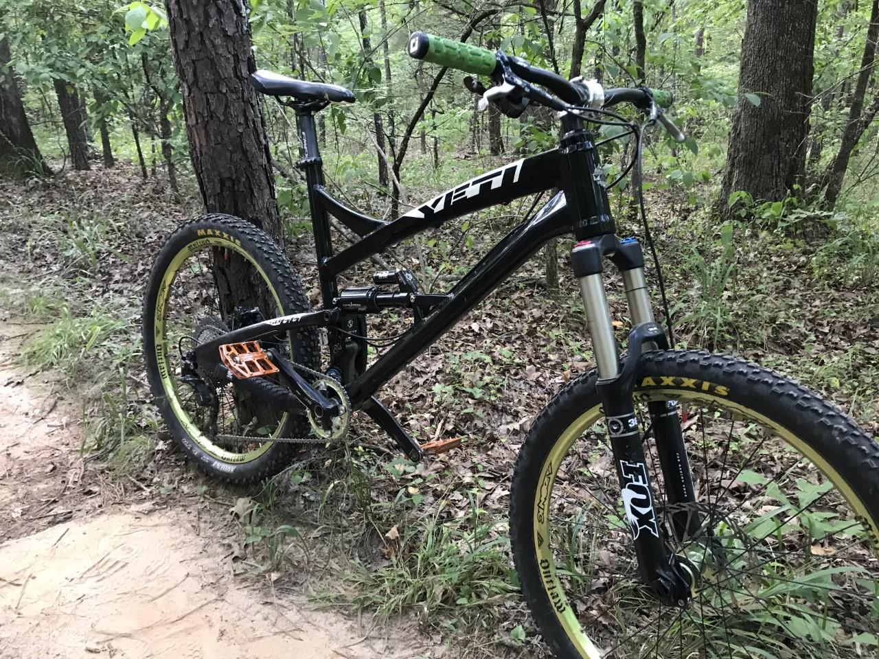 Yeti SB66: A mountain bike leaning against a tree on a woodland trail. The bike features a black frame, yellow-accented tires, and bright orange pedals. The surrounding area is lush with greenery and small plants, indicative of a natural, outdoor setting.