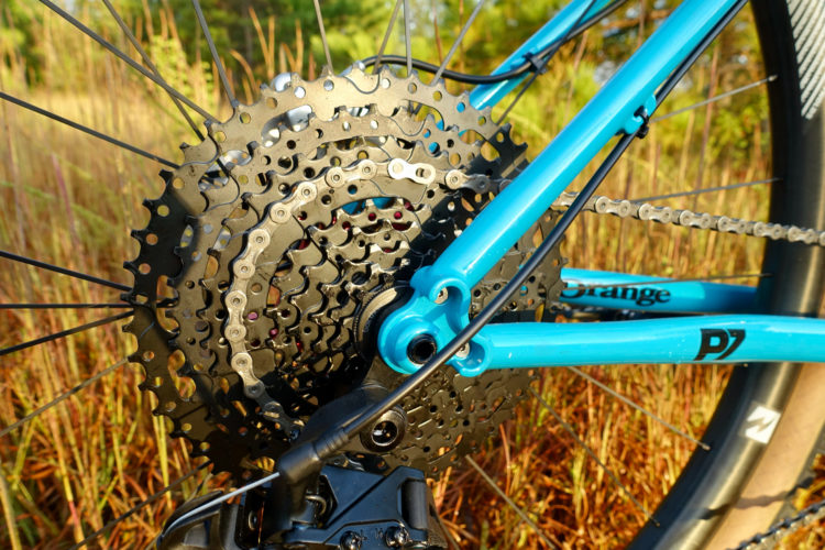 Close-up view of a bicycle's rear cassette and chain, featuring a vibrant blue frame and a background of tall grass. The image highlights the intricate gears and chain links designed for efficient cycling performance.