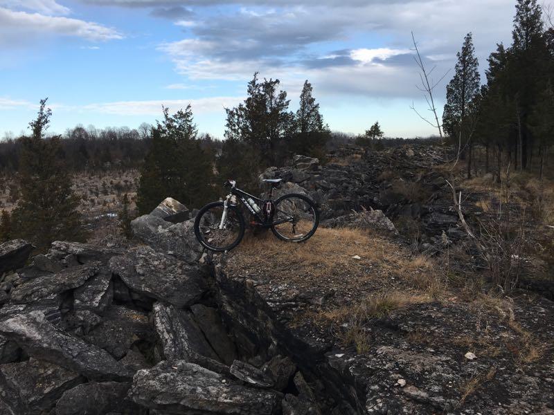 A mountain bike resting on rocky terrain, surrounded by sparse vegetation and trees under a cloudy sky. The landscape features a mix of rocky outcrops and open fields in the background, indicating a natural outdoor setting. East Quarry & North Shore Loops mountain bike trail.