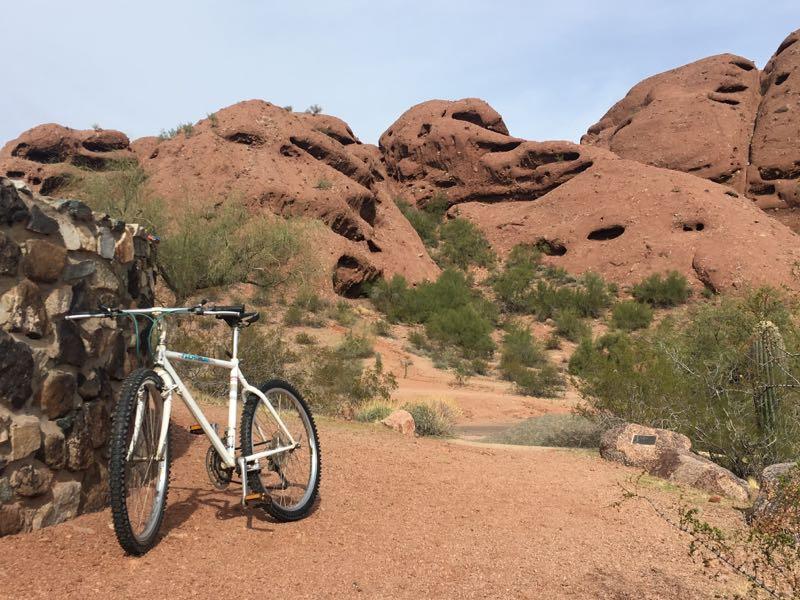 A white mountain bike stands next to a stone wall in a desert landscape, with reddish rock formations and sparse greenery in the background under a partly cloudy sky. Papago Park Area mountain bike trail.
