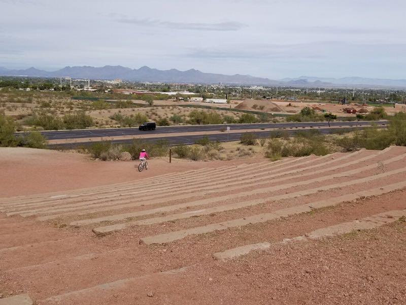 A cyclist in a pink shirt rides down a sloped trail made of concrete steps, with a backdrop of a desert landscape and distant mountains. A highway runs parallel to the trail, surrounded by sparse vegetation and buildings in the background under a mostly cloudy sky. Papago Park Area mountain bike trail.
