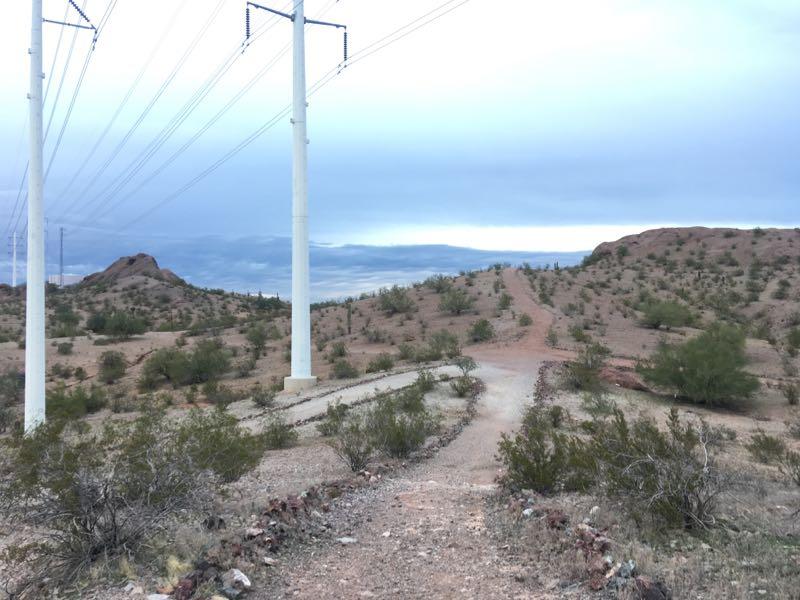 A dirt path winding through a desert landscape, bordered by sparse vegetation and hills in the distance. Power lines stretch across the sky above, set against a backdrop of overcast clouds. Papago Park Area mountain bike trail.