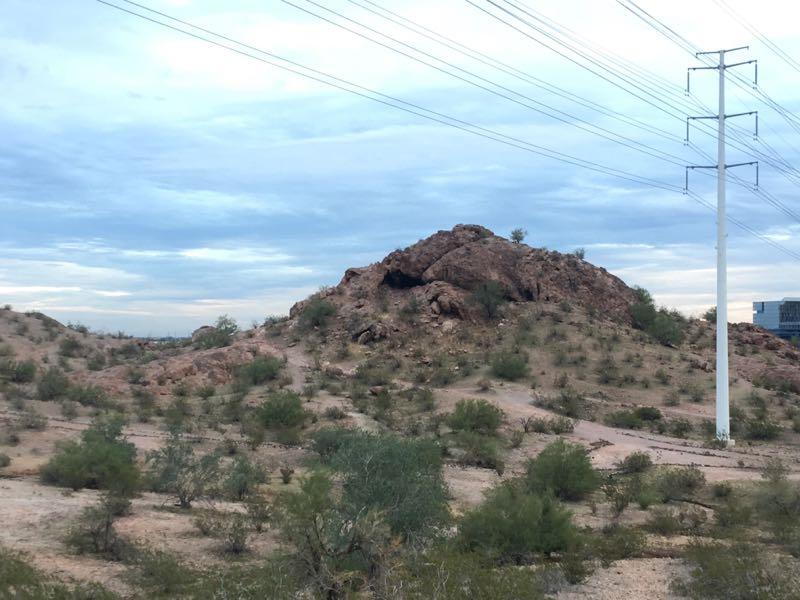 Rocky outcrop surrounded by sparse vegetation under a cloudy sky, with a power line running alongside it. Papago Park Area mountain bike trail.