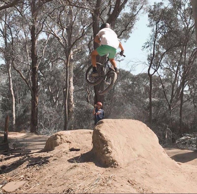 A person performing a bike jump over a dirt ramp in a wooded area, with trees in the background. Another individual stands nearby, observing the jump. The scene captures a moment of extreme sports excitement in a natural setting. Hudson