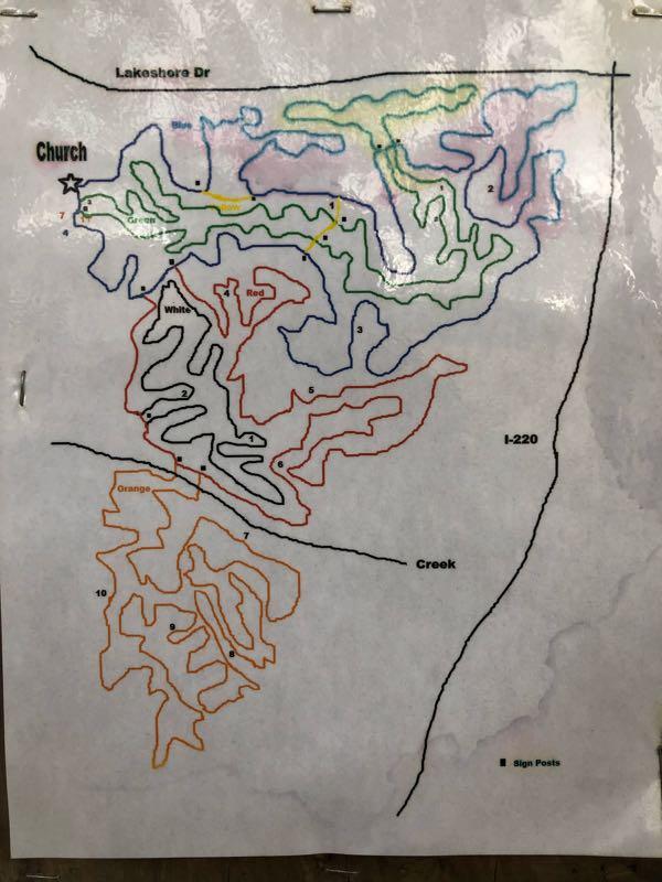 A hand-drawn map highlighting trails and paths, with colored lines indicating different routes. Key features include a labeled "Church" with a star icon, intersecting roads such as Lakeshore Drive and I-220, and distinct color-coded trails (blue, green, yellow, red, orange) along with numbered waypoints. Additional markings indicate the presence of sign posts and natural landmarks like a creek. The map is displayed on a sheet of paper with a rustic background. Lakeshore MTB Singletrack mountain bike trail.